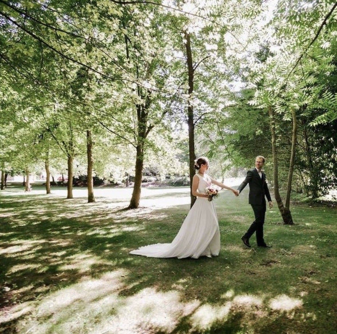 Couple de mariés se tenant la main dans le parc arboré du Domaine de la Cour des Lys, photographiés au cœur des arbres