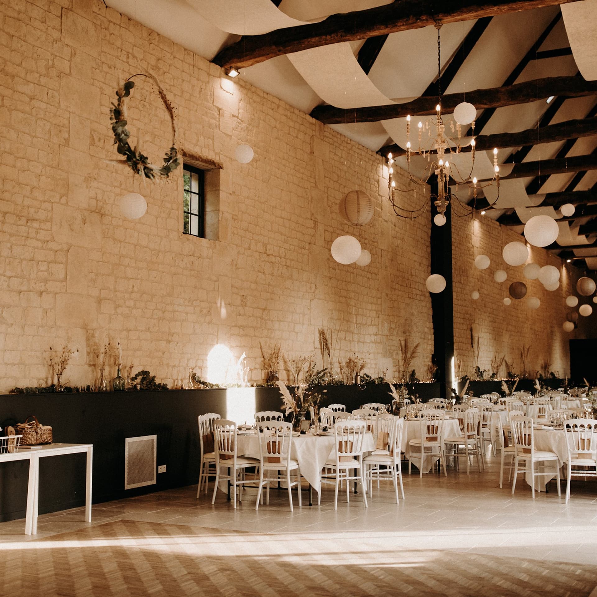 Grande salle de réception du Domaine de la Cour des Lys dressée pour un mariage — hauts murs en pierre de Caen, poutres anciennes, lustre et lampions au-dessus des tables rondes habillées de blanc