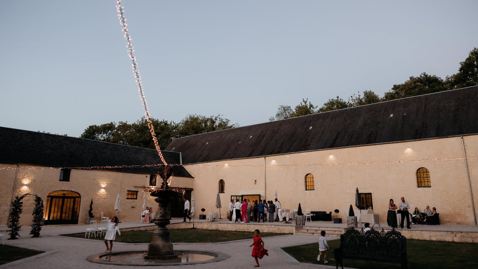 Cocktail de mariage au crépuscule dans la Cour d'Honneur du Domaine de la Cour des Lys — guirlandes lumineuses et invités autour de la fontaine, en Calvados