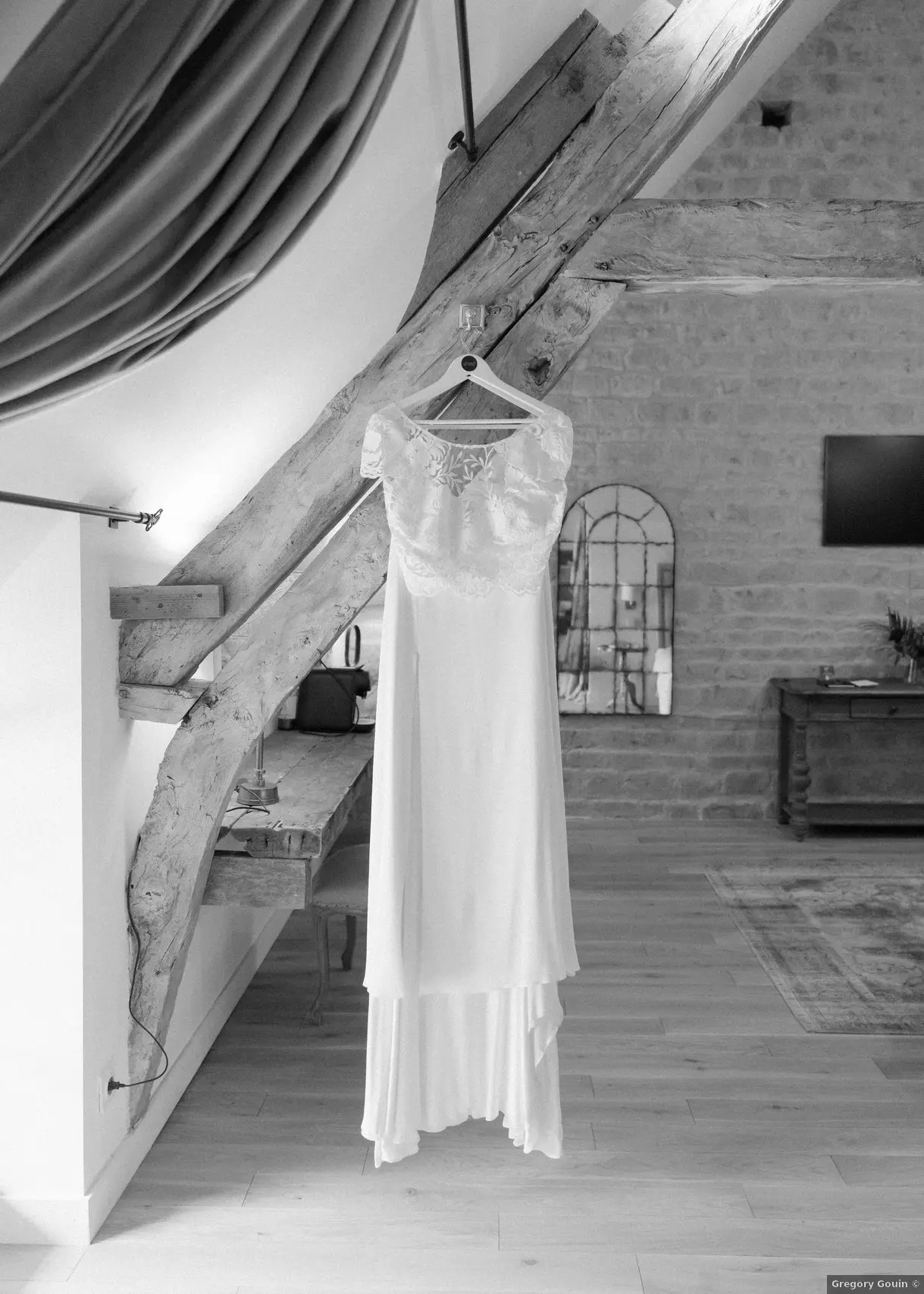 Préparatifs mariage Domaine de la Cour des Lys — robe Mademoiselle de Guise suspendue à une poutre ancienne XVIIᵉ siècle, photographie en noir et blanc