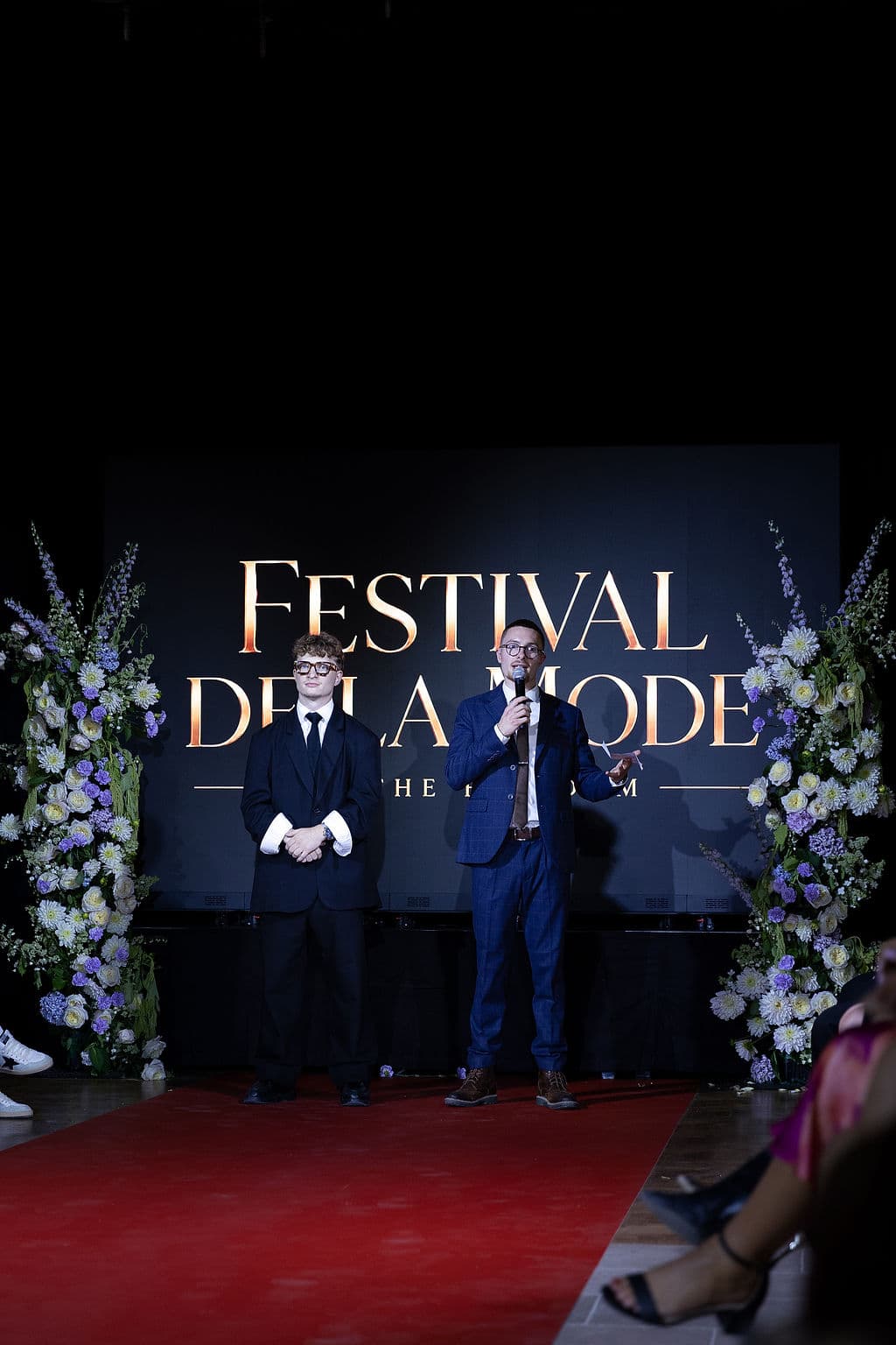 Clément Gouin et Esteban France présentent le premier Festival de la Mode de Caen sur la scène du Domaine de la Cour des Lys, devant le panneau lumineux et les compositions florales