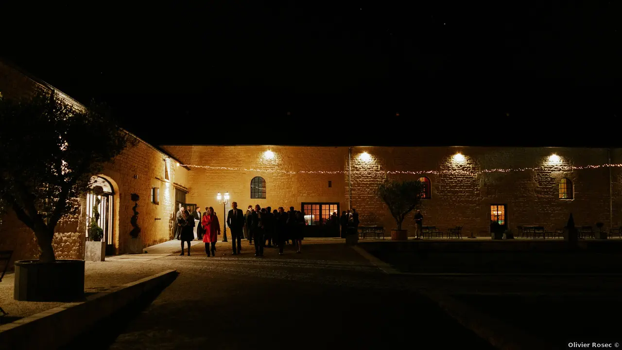 Cour d'honneur Domaine de la Cour des Lys au crépuscule — invités d'Adélaïde et Charles, guirlandes lumineuses, bâtisses en Pierre de Caen illuminées