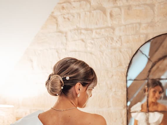 Louise se prépare le matin de son mariage — chignon orné de perles, mur en pierre apparente, reflet dans un miroir au Domaine de la Cour des Lys