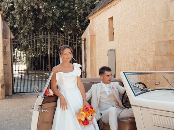 Louise et Baptiste assis dans une voiture vintage blanche devant le portail du Domaine de la Cour des Lys — bouquet aux couleurs vives
