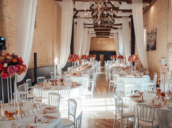 Grande salle de réception du Domaine de la Cour des Lys décorée pour le mariage de Louise et Baptiste — voilages blancs et compositions florales orange et rose